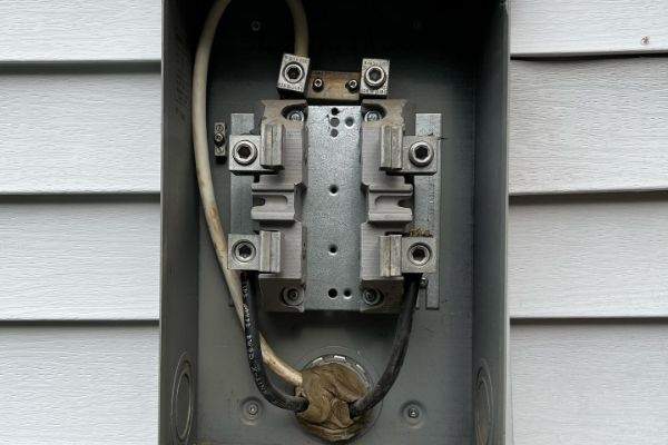 An empty electrical meter box with exposed wiring and terminals mounted on an exterior wall with gray siding.
