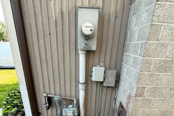 An electric meter and utility boxes mounted on the exterior wall of a building next to a gas meter and pipes.