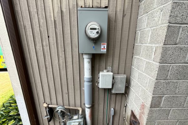 An electric meter box is mounted on the exterior wall of a building, with utility pipes and smaller connection boxes below it.