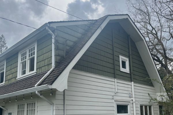 A two-story house with green and white siding, multiple windows, and a gambrel roof under a cloudy sky.