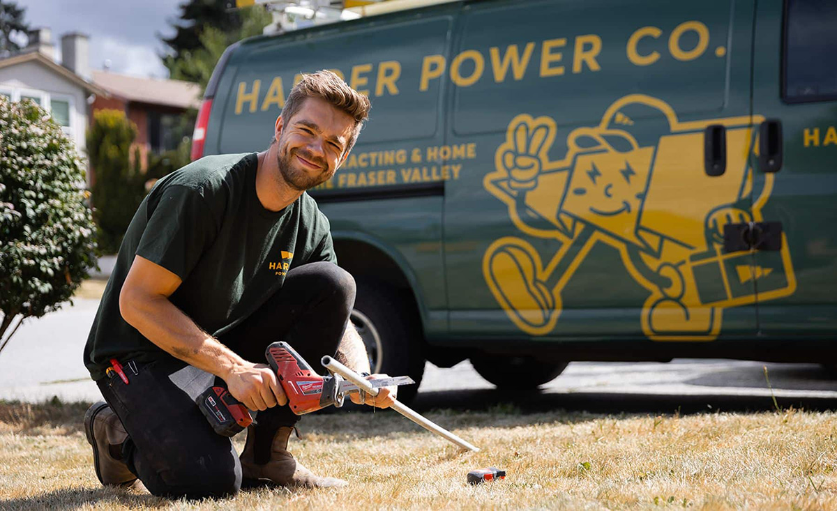 electrical-service-company-abbotsford A man kneels on grass holding a power drill, with a Harper Power Co. van displaying a cartoon mascot in the background.