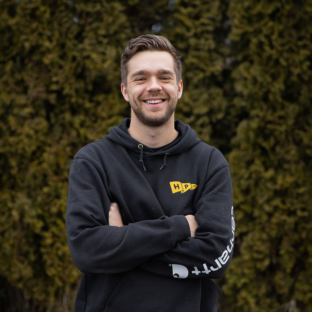A man in a black hoodie stands outdoors with his arms crossed, smiling at the camera. Green foliage forms the background.