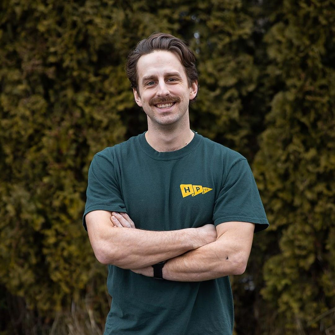 A man with brown hair and a mustache stands outdoors in front of green foliage, wearing a green T-shirt with a yellow "HPP" logo and his arms crossed.