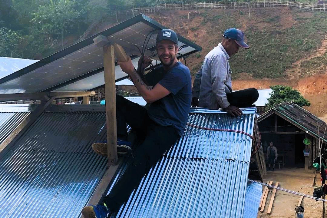 give-light-charity-program Two men install solar panels on a corrugated metal roof in a rural area, with one man smiling at the camera while working.