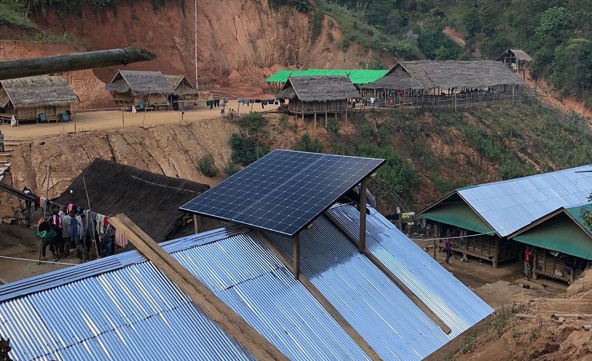 giving-light-charity-program-image-2 Solar panels installed on a metal roof in a rural village with thatched huts and dirt paths, surrounded by hills and greenery.