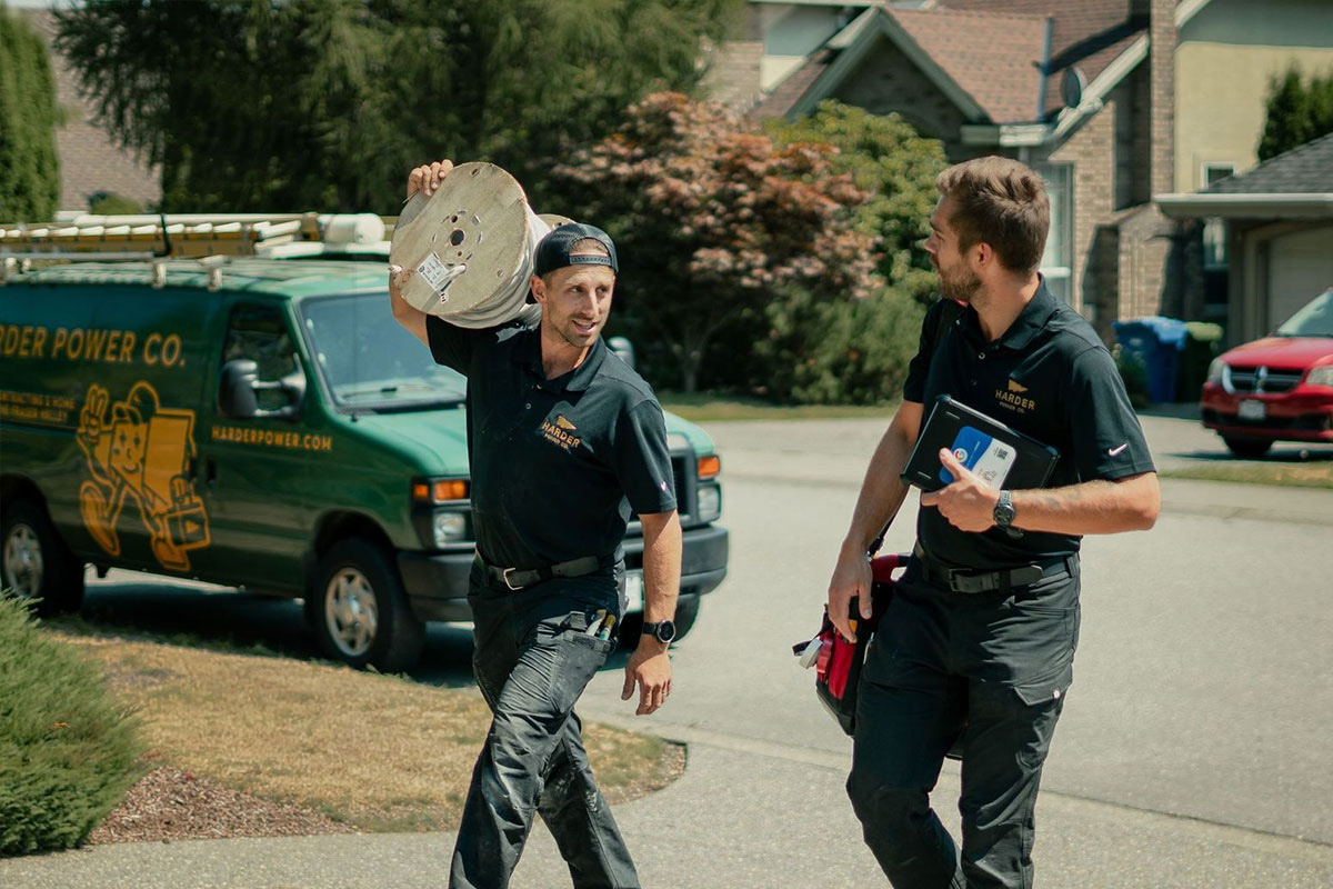 harder-power-electrical-abbotsford-bc Two electricians wearing work uniforms walk outside near a service van; one carries a large spool of cable while the other holds tools and paperwork.