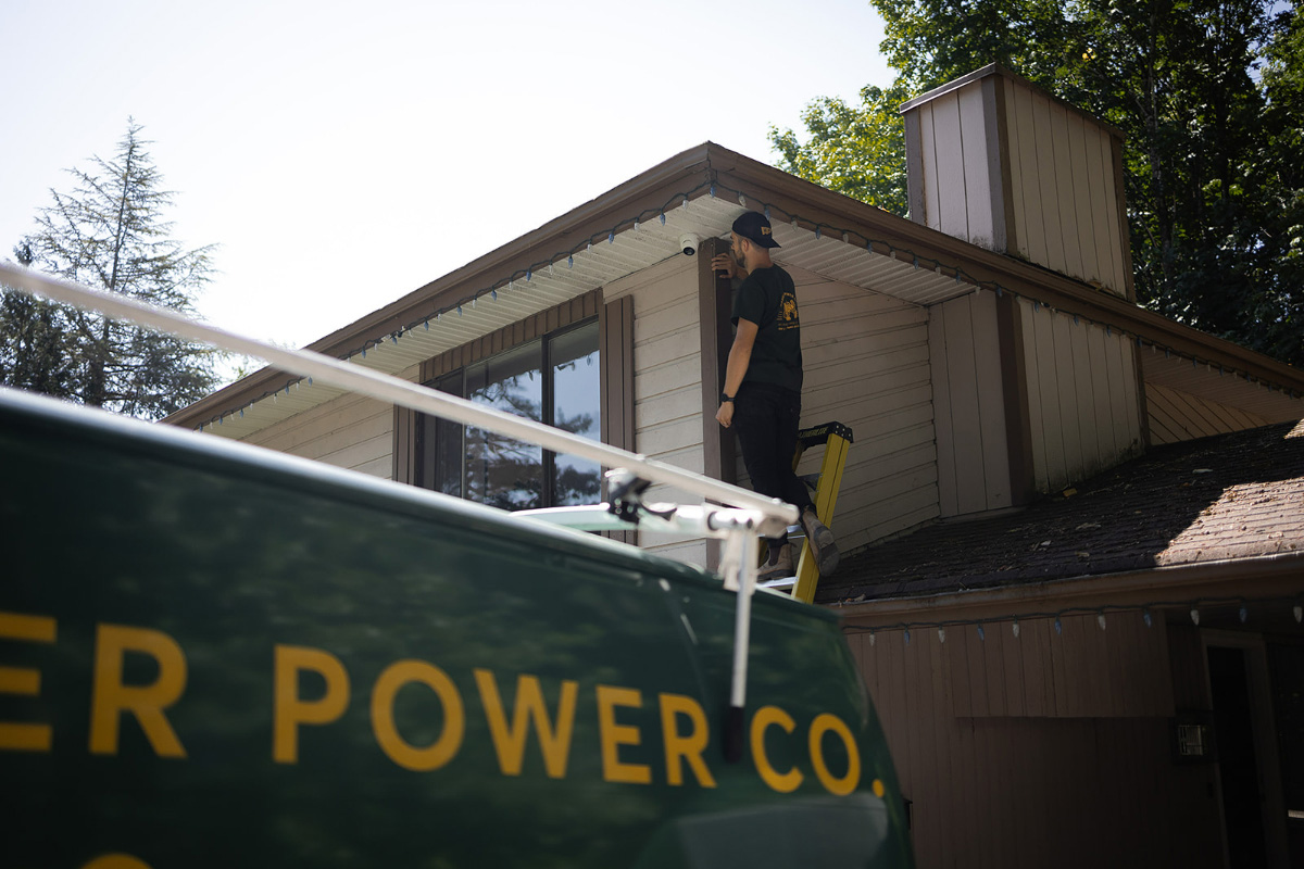 A worker on a ladder installs or inspects wiring along the roofline of a two-story house, with a green service van in the foreground.