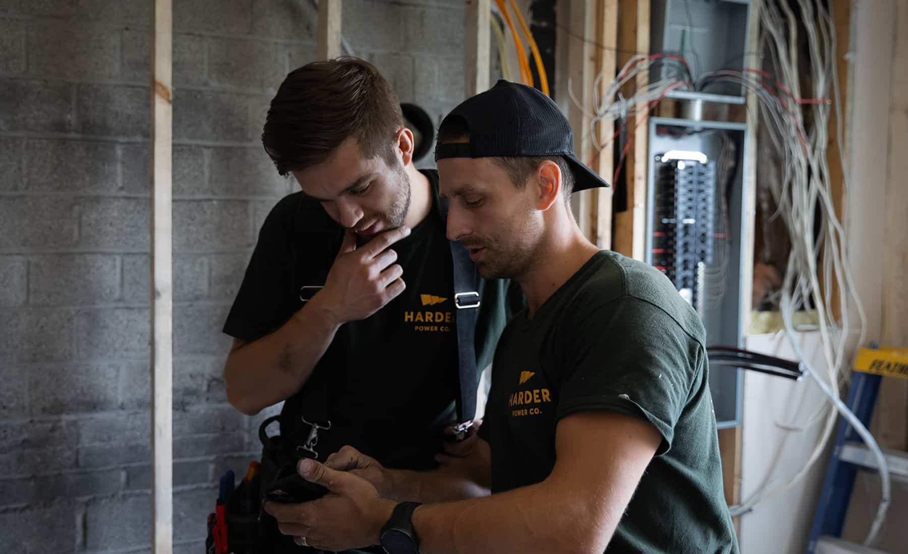 Two electricians in work attire examine a device together at a construction site with exposed wiring and a circuit breaker panel in the background.