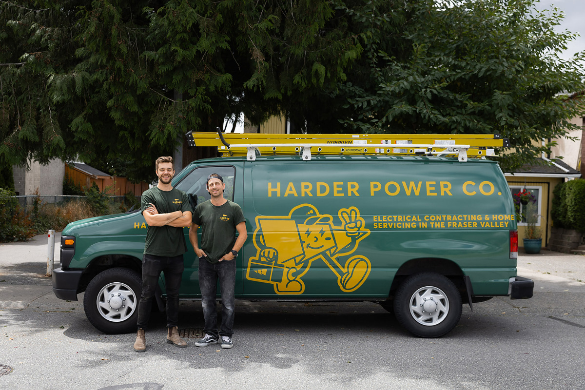 Two men in green shirts stand in front of a green Harder Power Co. van with a yellow ladder on top, parked on a residential street.
