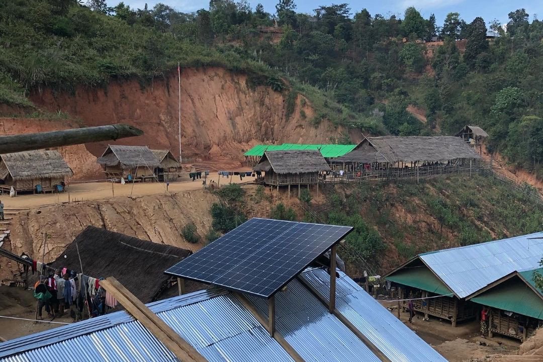 A rural village with thatched huts and tin roofs, some topped with solar panels, set on a hillside with exposed reddish soil and surrounding greenery.