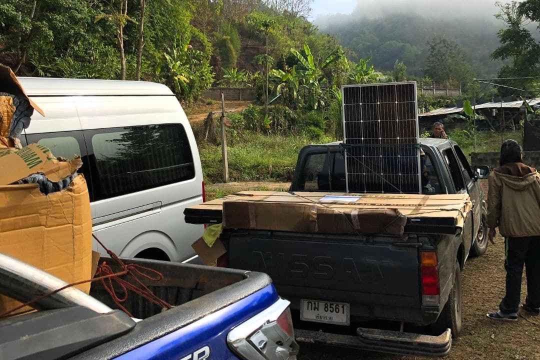 A pickup truck loaded with solar panels and boxes is parked next to a white van in a rural, green area with trees and hills in the background.