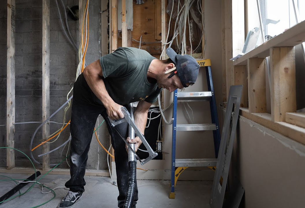 A person in work clothes cuts a flexible metal conduit in a room under renovation, with exposed wiring, a ladder, and construction materials visible.