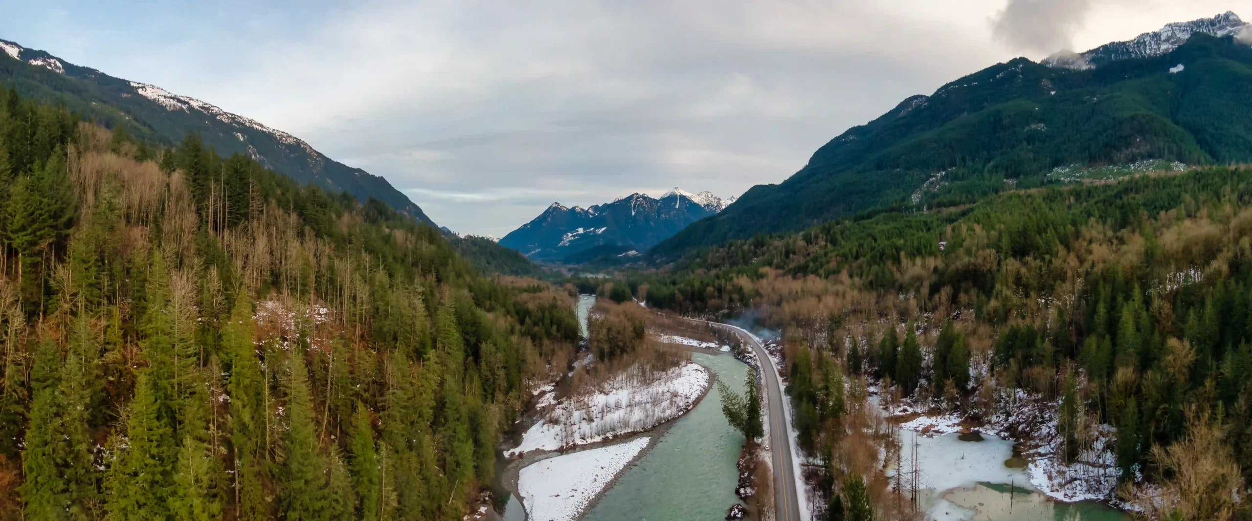 A river runs alongside a road through a forested valley, with snow patches on the ground and mountains in the background under a cloudy sky.