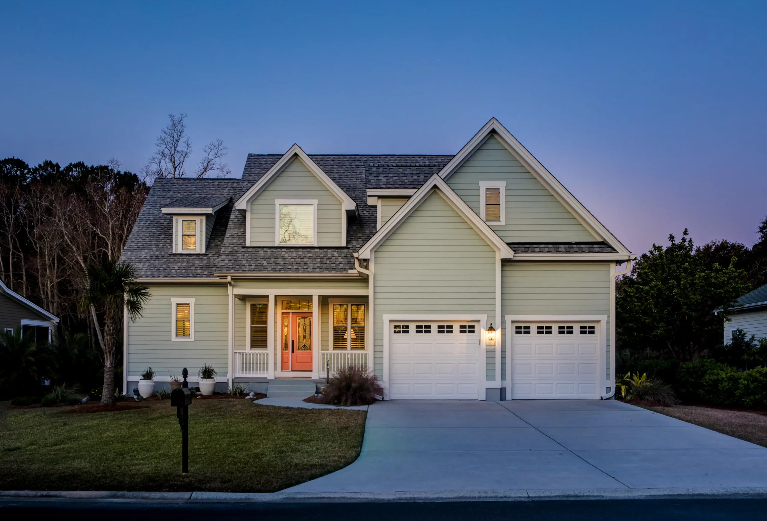 Two-story suburban house with light green siding, large front porch, and double garage, photographed at dusk with lights on inside.