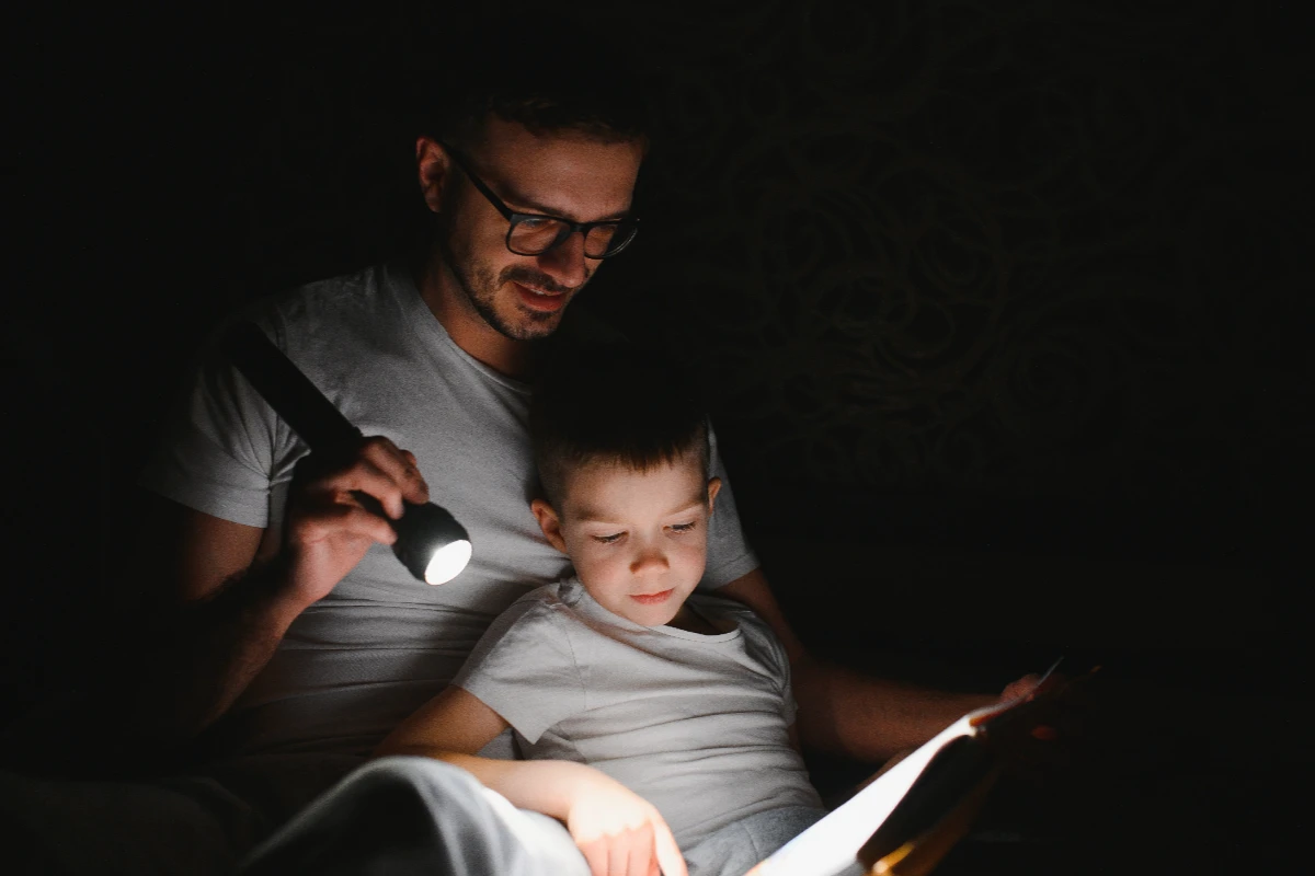 An adult and a child sit in the dark, reading a book together with a flashlight illuminating the pages.