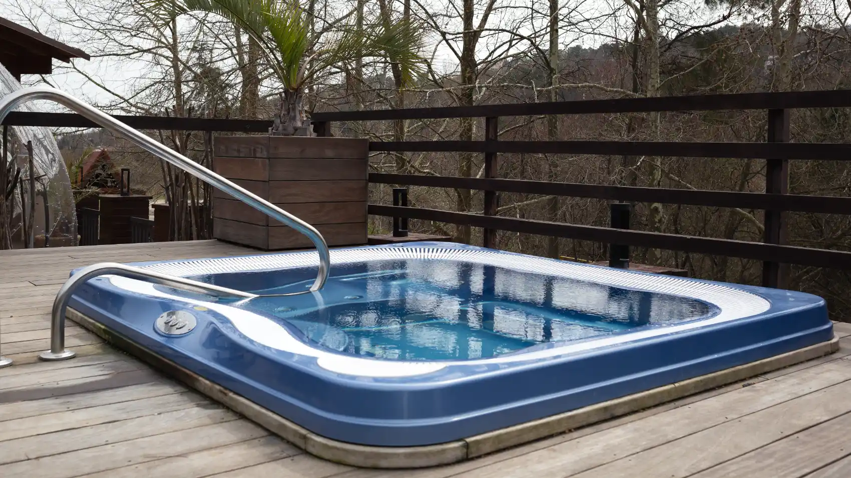 A blue hot tub is installed on a wooden deck with a metal handrail, perfect for relaxing in Fraser Valley homes, surrounded by a railing and trees in the background.