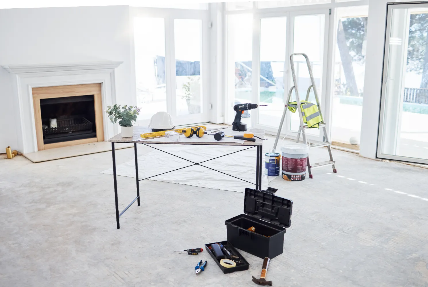 A room under renovation in a Fraser Valley home features a table holding tools, a toolbox on the floor, a ladder, paint cans, a hard hat, and an electrical panel upgrade near the fireplace in the background.
